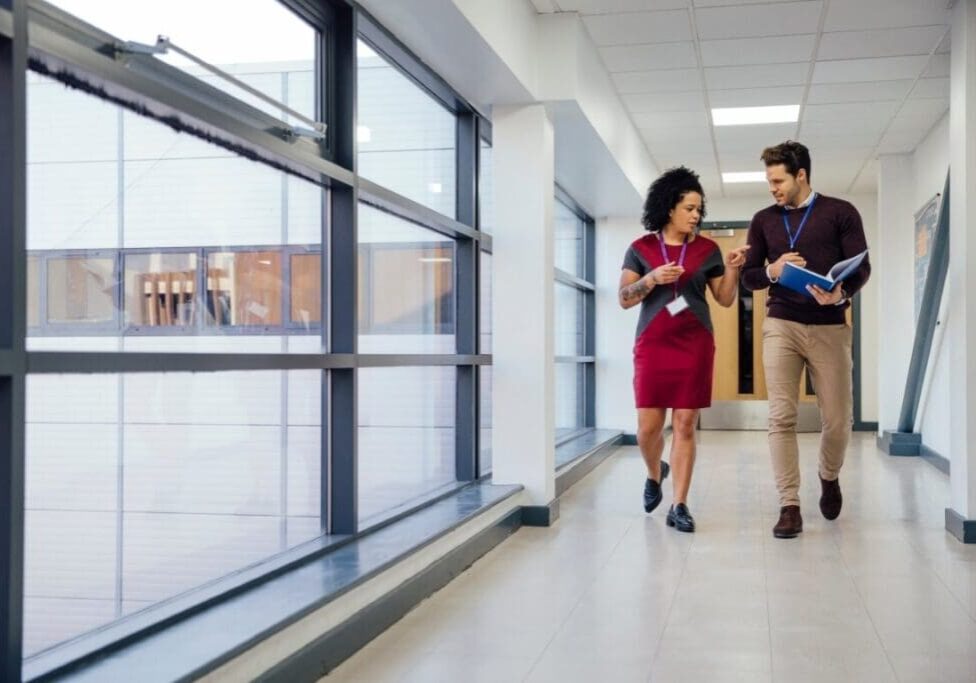 Two people walking in a hallway near some windows.